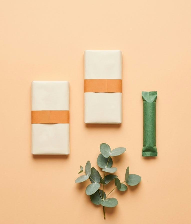 Plastic-free pads and a tampon in paper wrappers on a warm peach background — the products COVERED dispenses free